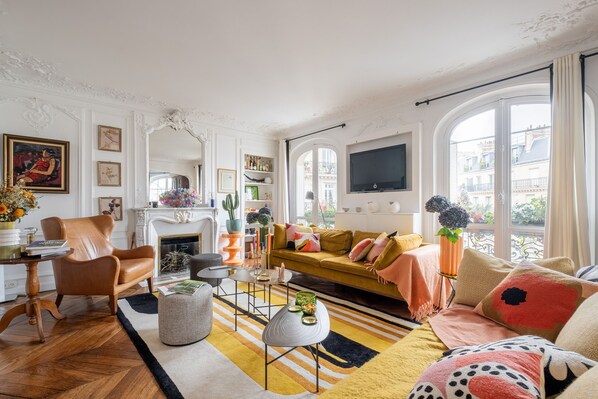 A well-lit living room with a sofa, armchair, and coffee table, featuring a fireplace, large windows, and decorative items. - Sunrise Synergy, by Veeve (Paris)