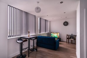 A modern living room with a blue sofa, a dining table with chairs, a TV, and a large window with blinds.