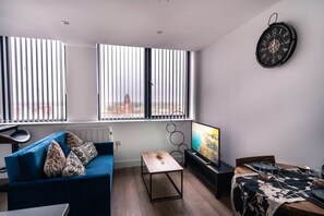 A modern living room with a blue sofa, a small table, a TV, and a dining area with a table set for two.