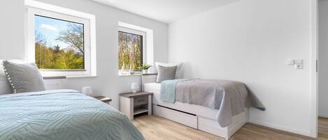 A modern bedroom with two beds, a nightstand, and a window with a view of trees.