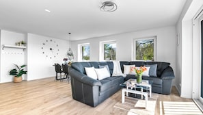 Uma sala de estar moderna com um sofá modular cinza escuro, uma mesa de centro de vidro e um tapete branco. Duas janelas oferecem luz natural e um relógio de parede é visível.
