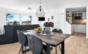 Uma sala de estar moderna com um sofá de cor escura, uma mesa de jantar com cadeiras e uma área de cozinha com armários brancos e bancada de madeira.