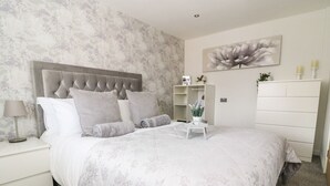 Une chambre à coucher avec une tête de lit capitonnée, une couvre-lit à motifs floraux, une table de chevet avec une lampe, une commode et un grand tableau floral accroché au mur.