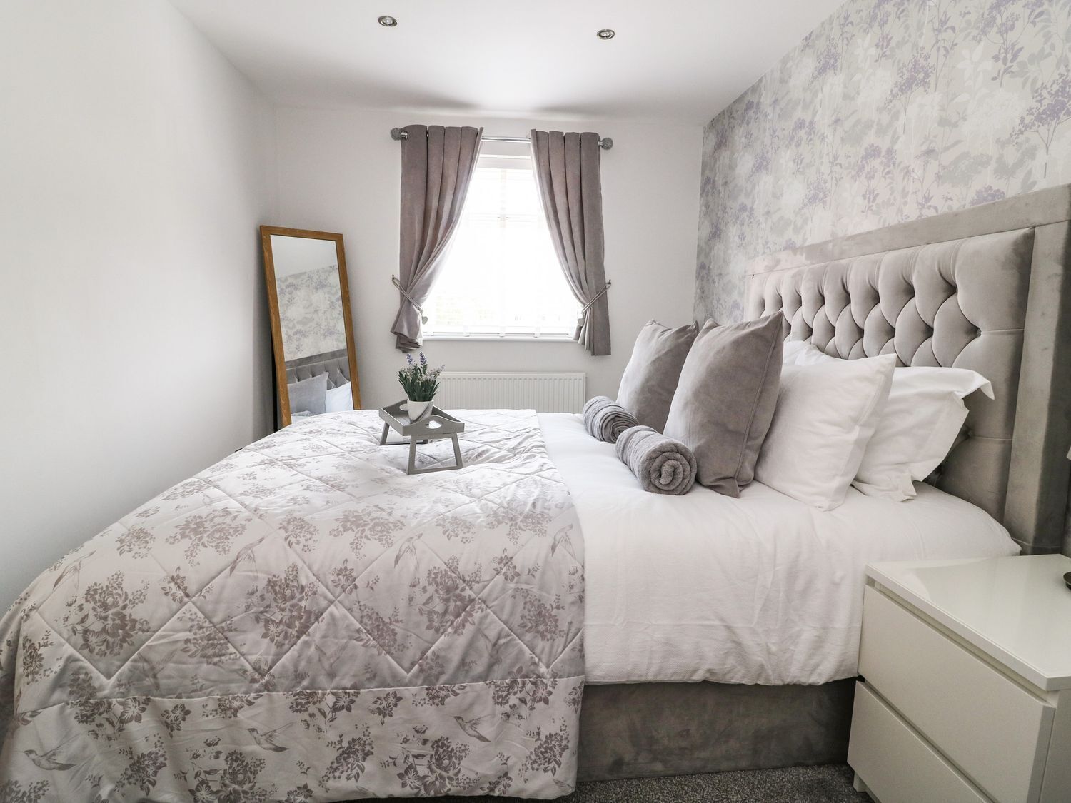 A bedroom with a large bed, a bedside table, a nightstand, a window with curtains, and a floral wallpaper.