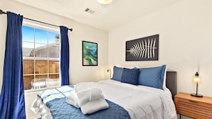 Une chambre à coucher avec un grand lit, une fenêtre avec des rideaux, une table de chevet avec une lampe et deux œuvres d’art murales.