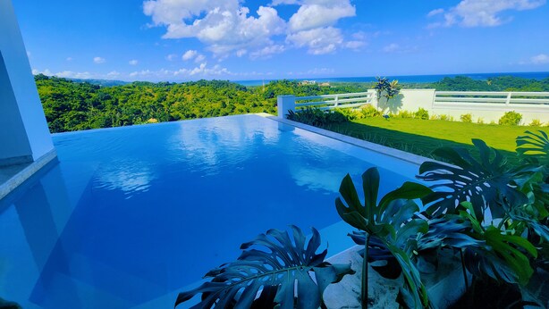 Une piscine à débordement moderne offrant une vue dégagée sur un paysage verdoyant et luxuriant, ainsi que sur l’océan à l’horizon.
