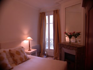 A hotel room with a bed, a nightstand with a lamp, a window with curtains, and a mirror above a fireplace. - Central Parisian apartment. A 5 minute walk to the Notre-Dame and the Seine (Paris)