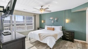 Une chambre à coucher comprenant un lit, des tables de chevet, un bureau, un ventilateur de plafond et une vue sur la ville grâce à de grandes fenêtres.