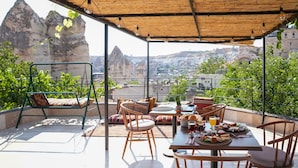 A terrace with a table set for breakfast, chairs, and a swing chair, overlooking a mountainous landscape. - Matiana View Cave Hotel (Nevsehir)
