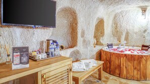 A room with a wooden table, a large flat-screen TV, a wooden bathtub, and a stone wall. - Matiana View Cave Hotel (Nevsehir)