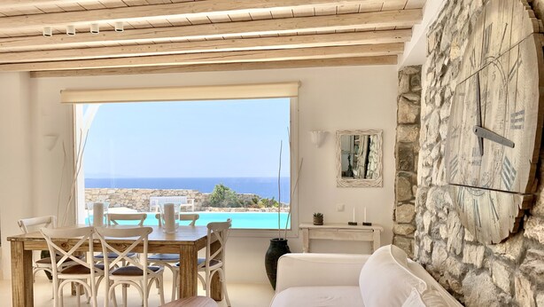 Ein Wohnzimmer mit Steinwand, Holzboden, einem weißen Sofa, einem Esstisch mit Stühlen und Meerblick.