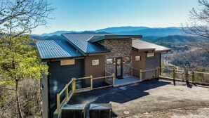 A modern cabin with a metal roof, stone walls, and a wooden deck. It features a staircase leading to a door and a view of mountains in the background. - Infinity View Lodge - Indoor pool & mountain views (Sevierville)
