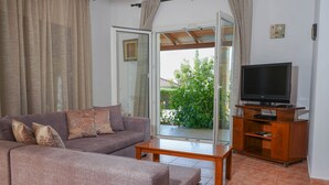 Ein Wohnzimmer mit einer Couch, einem Couchtisch, einem Fernseher auf einem Holzgestell und einer Glastür, die in einen Garten führt.
