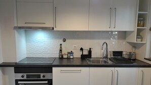 A modern kitchen with white cabinets, a black countertop, a built-in oven, a sink, and various kitchen utensils. - Apartamento Acolhedor em Faro, a Poucos Minutos do Centro e do Barco (Faro)