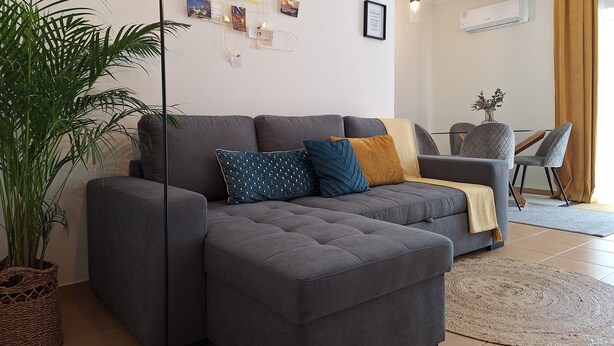 A modern living room with a grey sectional sofa, a floor lamp, a wall-mounted photo display, and a dining area with a chair and table. - Apartamento Acolhedor em Faro, a Poucos Minutos do Centro e do Barco (Faro)