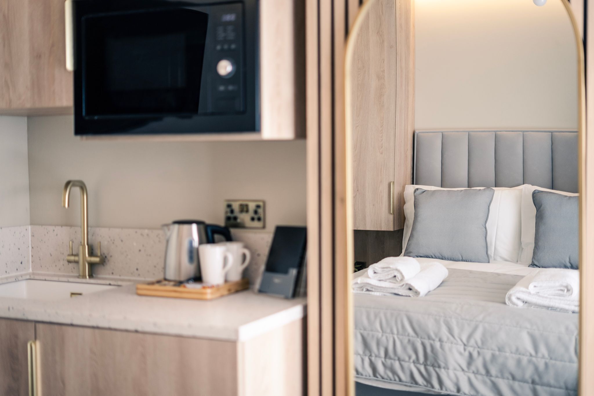 Habitación de hotel compacta con kitchenette, que incluye microondas, pileta y pequeño espacio para el desayuno. La habitación tiene una cama con ropa de cama gris y un cabecero, y un marco de puerta de madera.