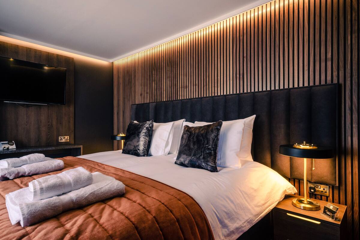 Una habitación de hotel moderna con una cama grande, un televisor de pantalla plana montado en la pared, una mesita de noche con una lámpara y toallas cuidadosamente dobladas.