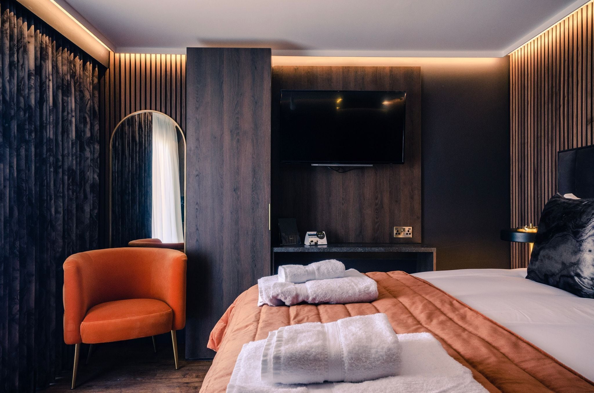 Habitación de hotel moderna con una cama grande, un televisor de pantalla plana montado en la pared, un panel de pared de madera y una sola silla naranja.