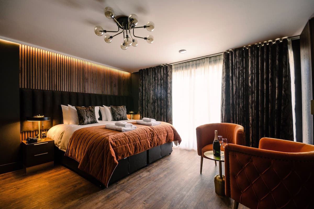 Habitación de hotel moderna con una cama grande, una mesita de noche con lámpara, una silla y una mesa con una botella y vasos.
