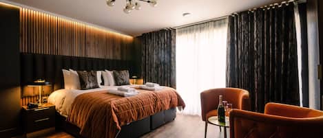 A modern hotel room with a large bed, a nightstand with a lamp, a chair, and a table with a bottle and glasses.