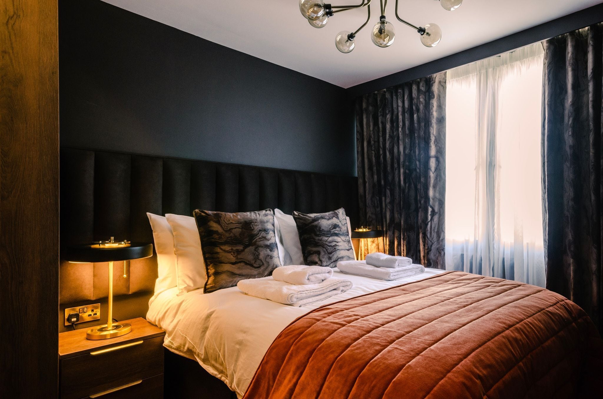 A modern hotel room with a large bed, dark walls, and a view of the city through sheer curtains.