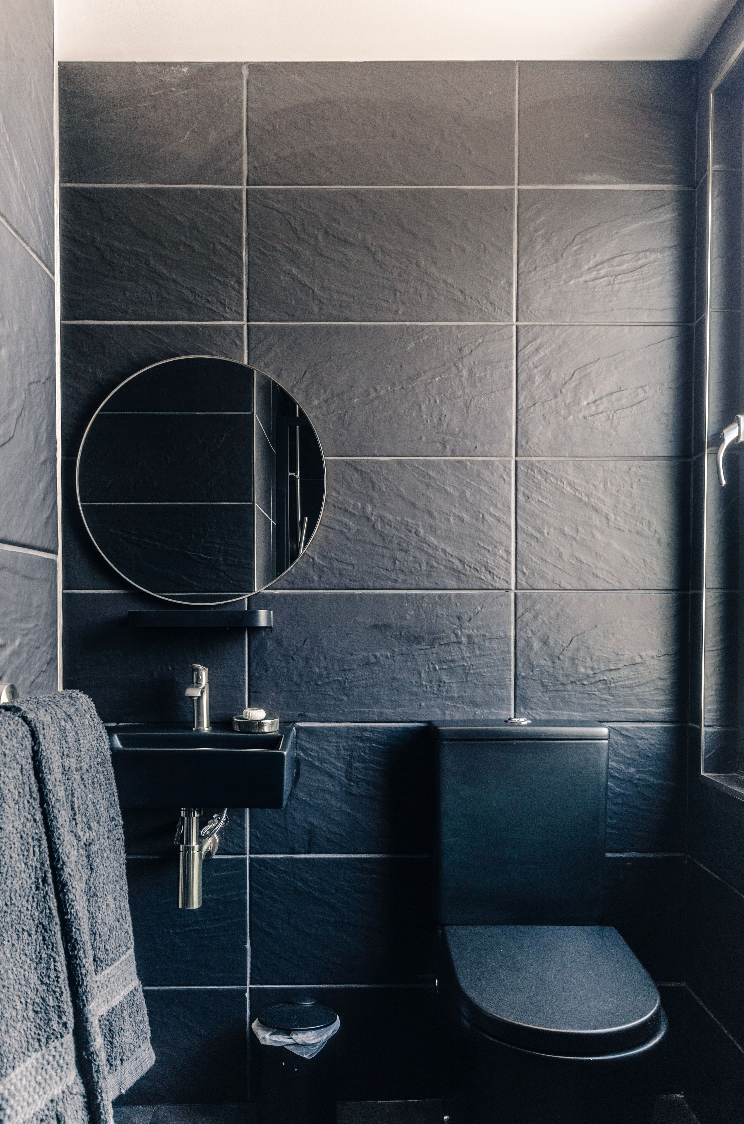 A modern bathroom with dark tiles, a round mirror, a sink, and a toilet.