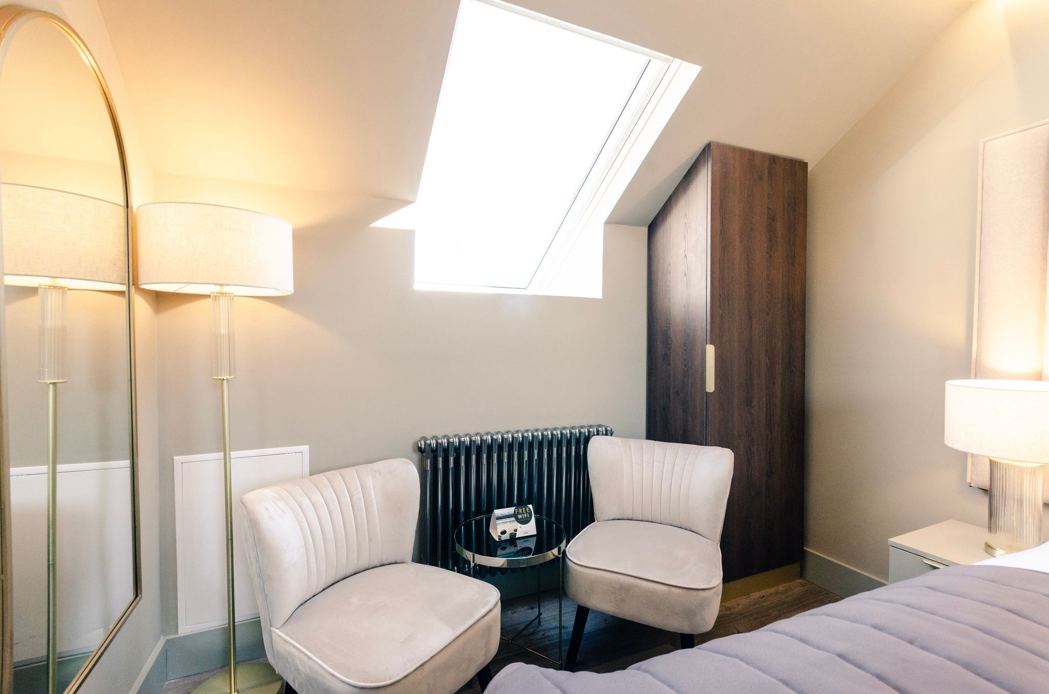 Une chambre d’hôtel avec une lucarne, deux fauteuils, une petite table ronde, un lit, un radiateur et une armoire.