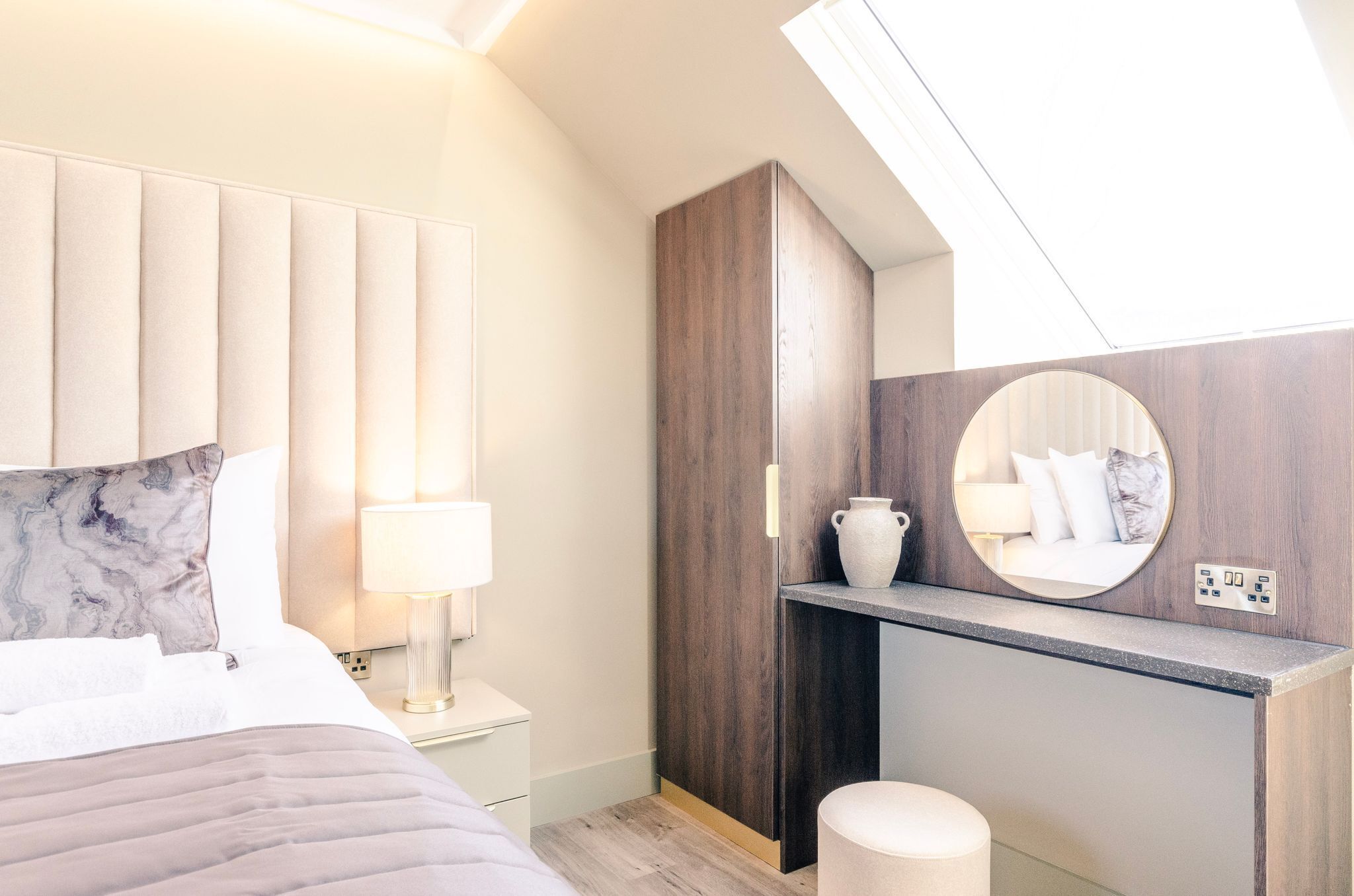 A modern bedroom with a bed, bedside table, lamp, wooden wardrobe, and a round mirror on the wall.