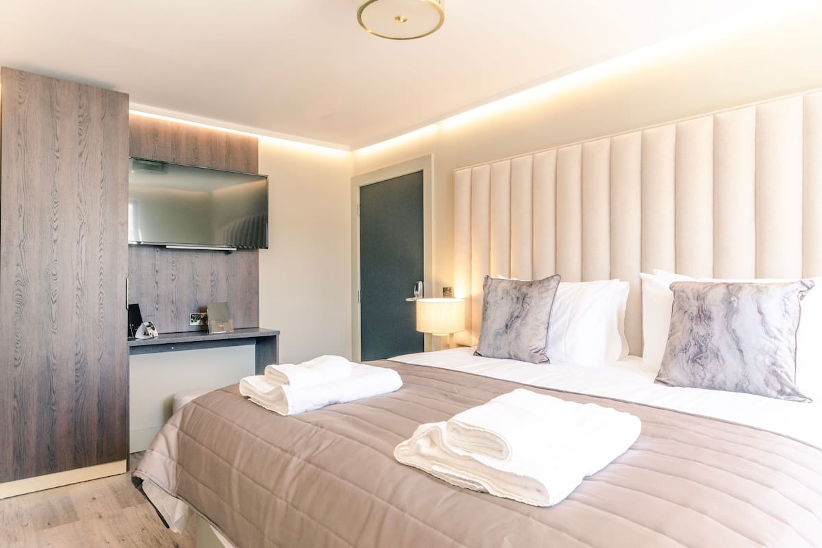 Habitación de hotel moderna con una cama grande, un televisor de pantalla plana montado en un panel de madera, una mesita de noche con lámpara y un cabecero con paneles verticales.