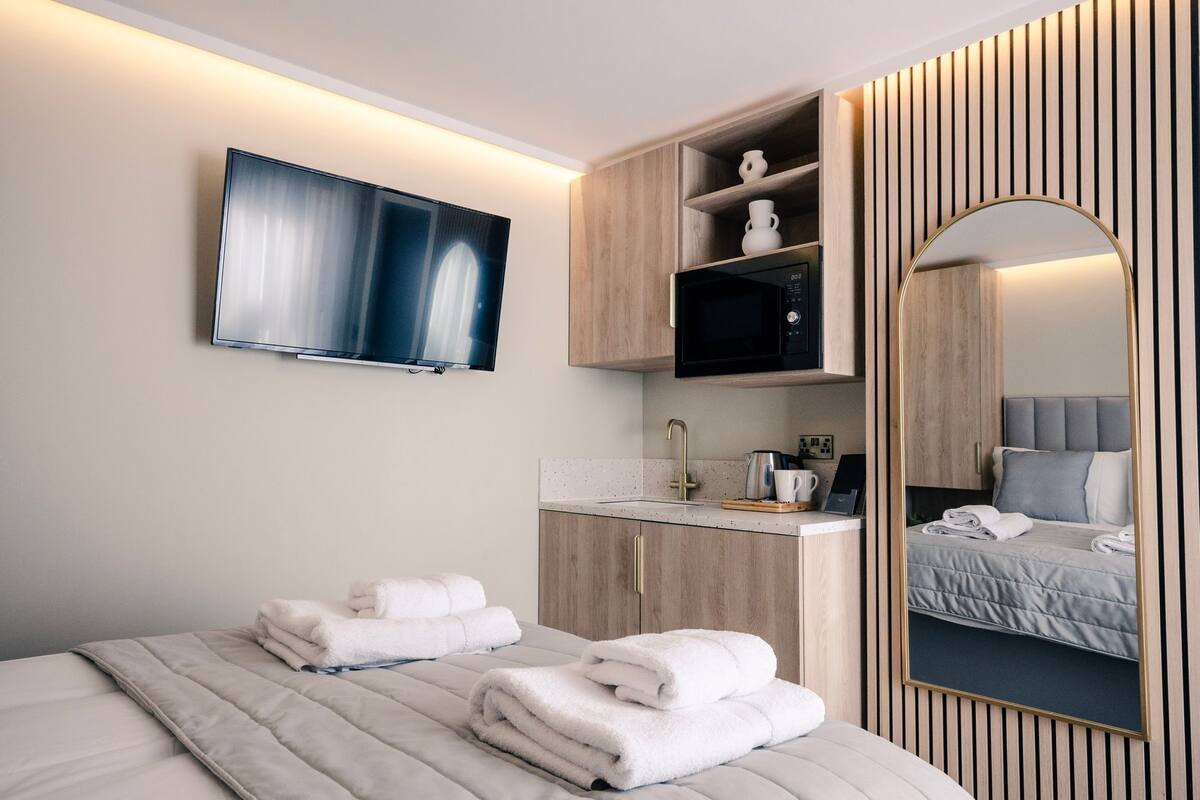 Una habitación de hotel moderna con un televisor de pantalla plana montado en la pared, un tocador de madera con un lavamanos y un espejo grande que refleja la cama.