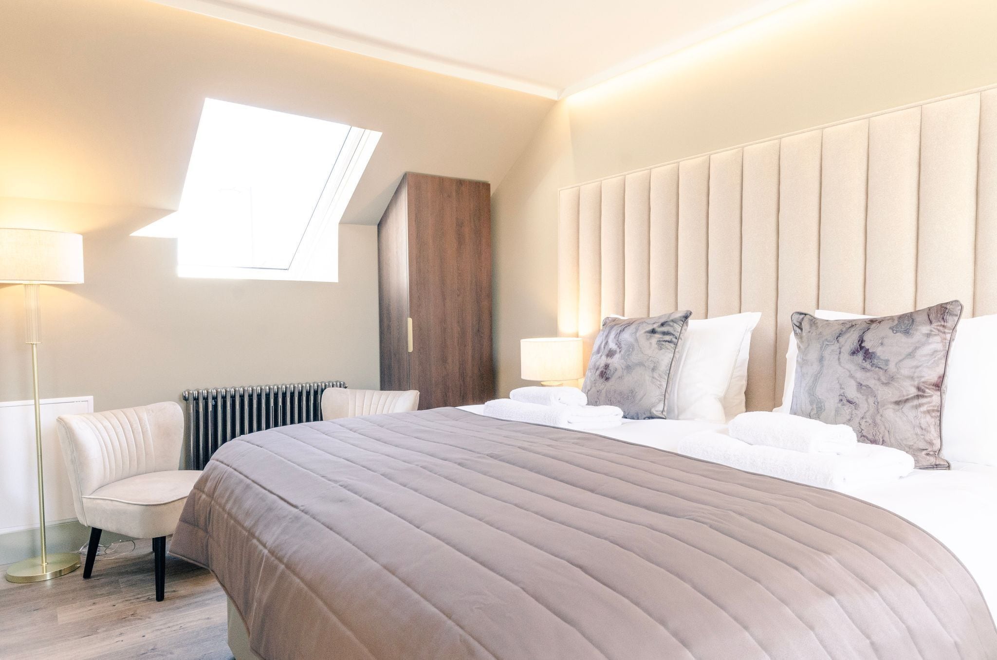 A modern hotel room with a large bed, a headboard, a radiator, a chair, and a floor lamp.