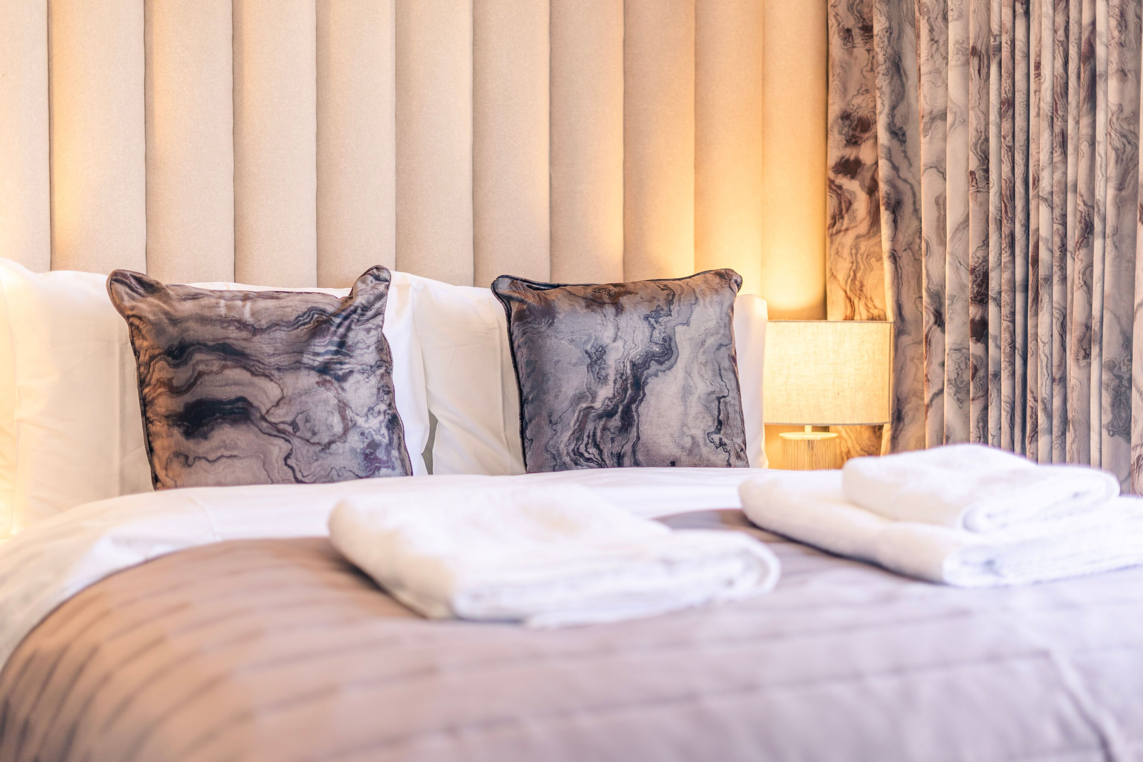 A neatly made bed with two pillows, a bedside lamp, and a window with curtains.