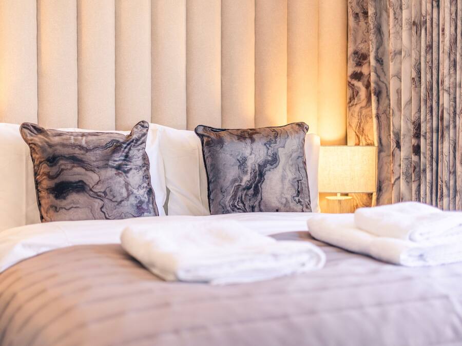 Una cama bien hecha con dos almohadas, una lámpara de noche y una ventana con cortinas.