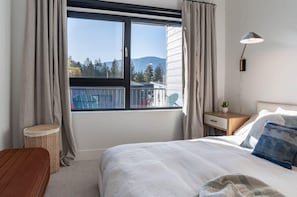 Une chambre avec une grande fenêtre, un lit, une table de chevet, une lampe et une vue sur un immeuble et des arbres.