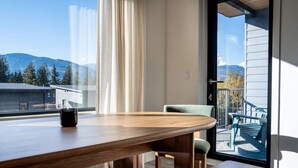 A modern dining area with a wooden table, a chair, and a view of mountains through a large window. - Snowed Inn - Luxurious 3 Bedroom Condo (Revelstoke)