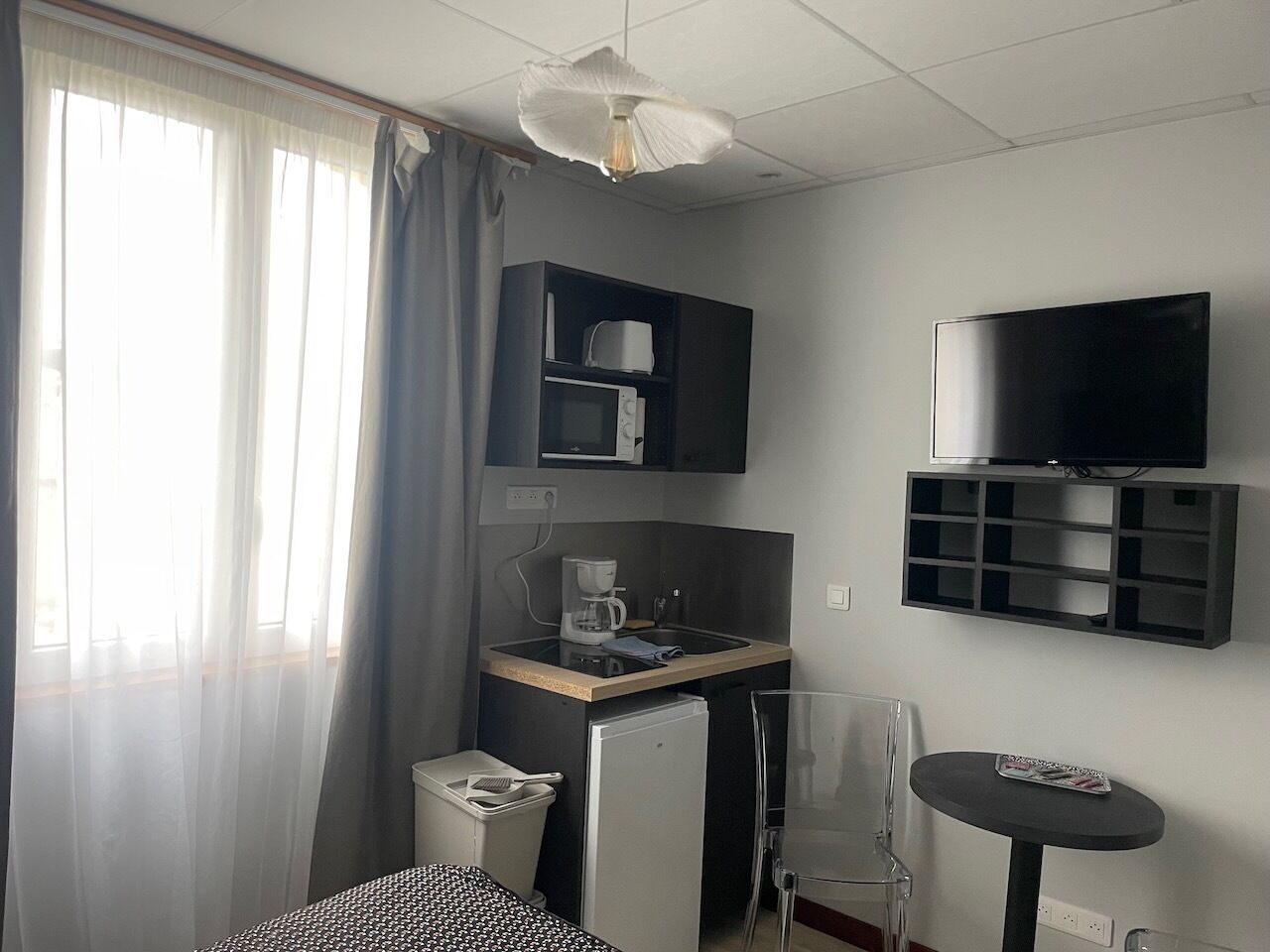 Ein kompaktes Hotelzimmer mit Küchenzeile, Flachbildfernseher, kleinem Esstisch und einem Fenster mit durchsichtigen Vorhängen.