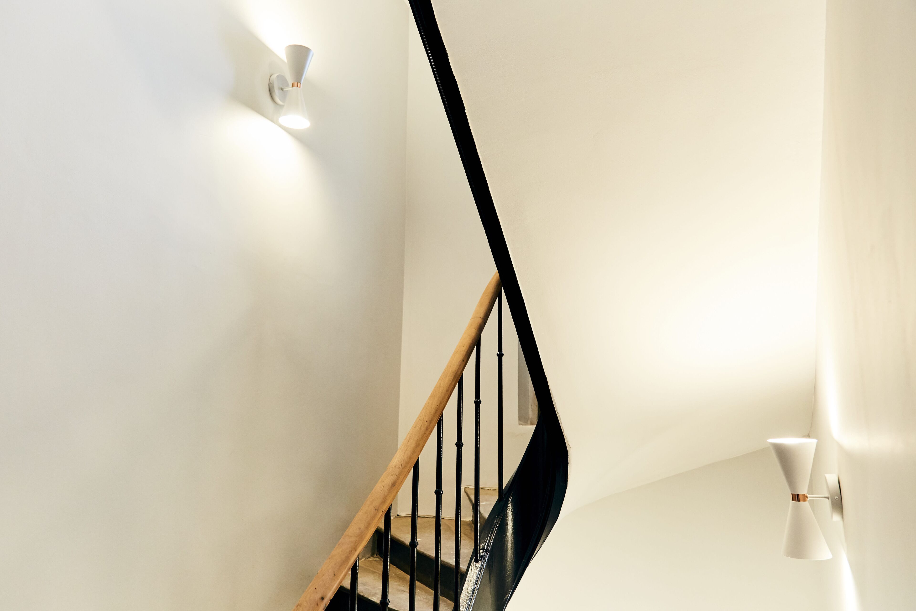 A modern staircase with a wooden handrail and black balusters, leading to a landing with a white wall and a wall-mounted light fixture.