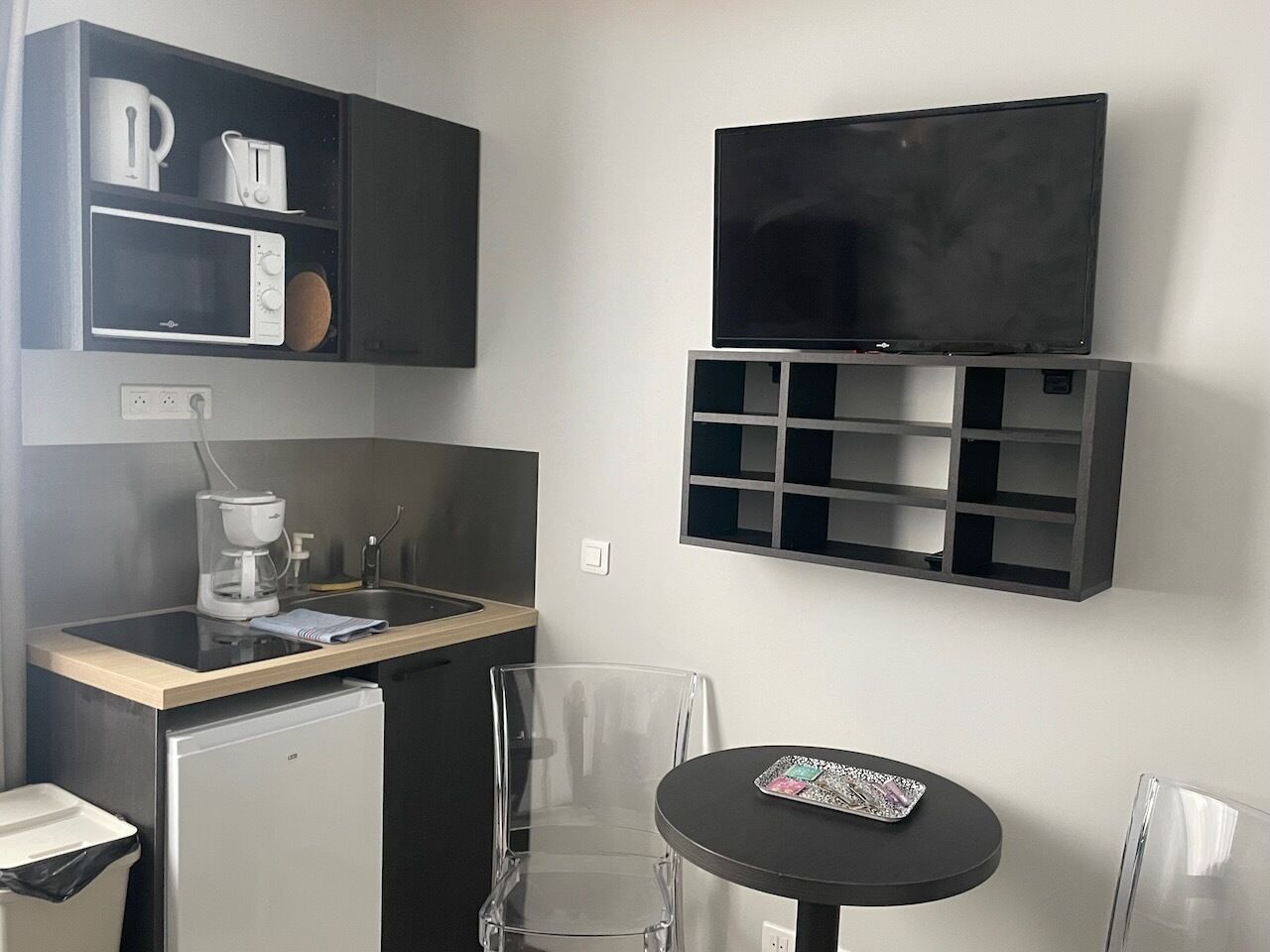 Una kitchenette compacta con microondas, cafetera y refrigerador pequeño. Hay un televisor montado sobre una unidad de estanterías, y una mesa con una bandeja.