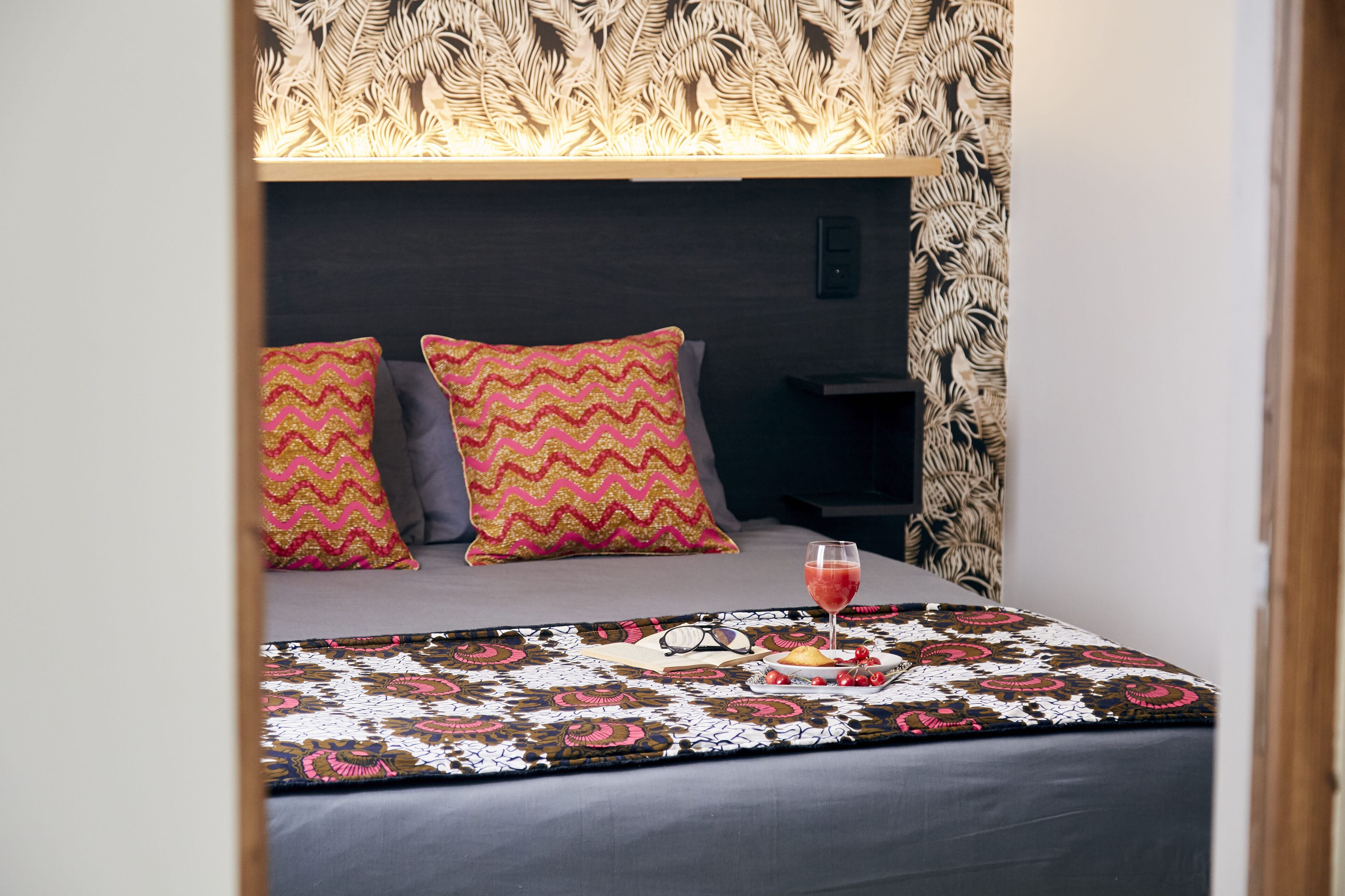 Una cama bien hecha con almohadillas estampadas, una mesita de noche y una bandeja con bebidas y aperitivos.