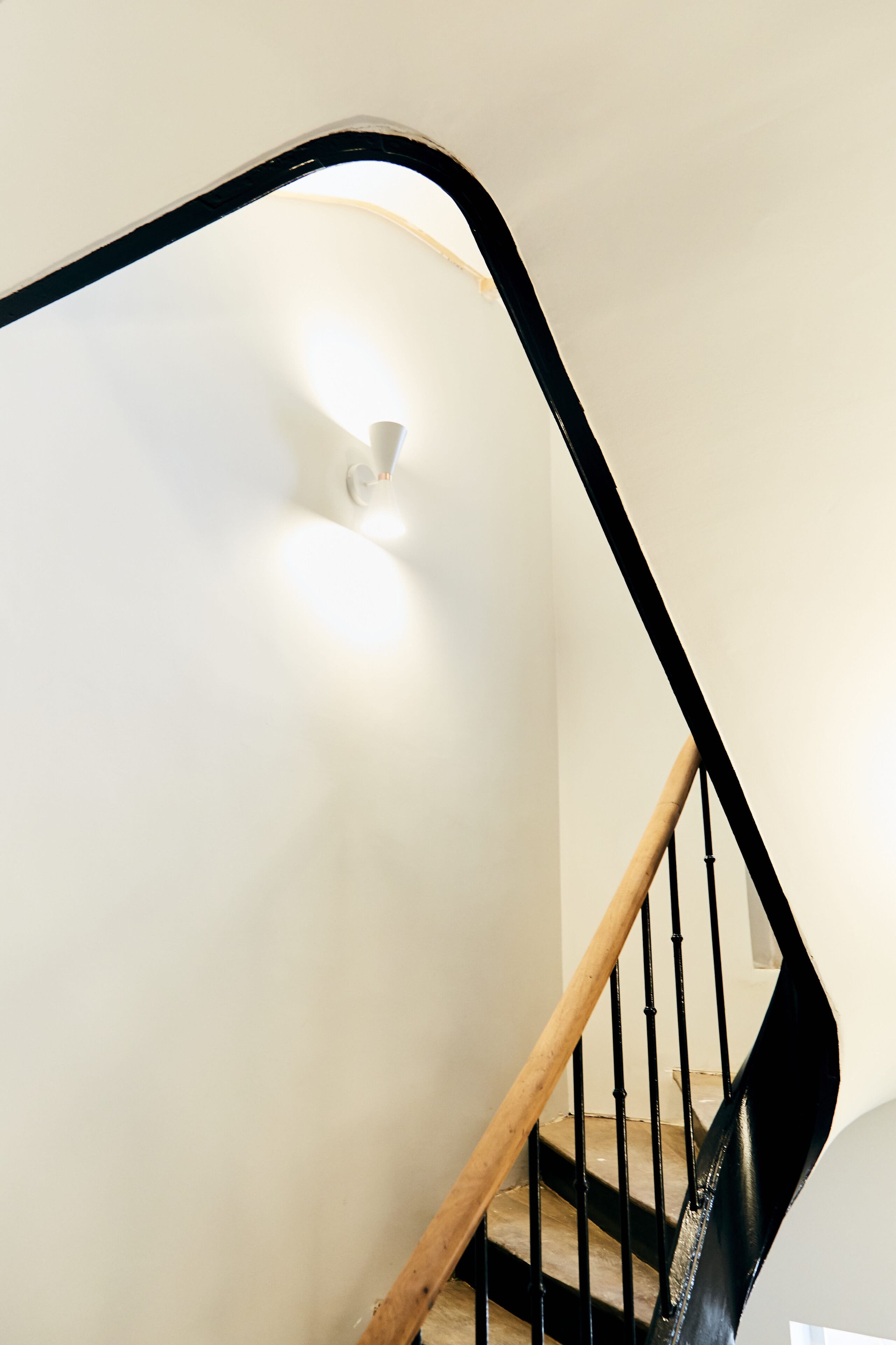A modern staircase with a wooden handrail and black balusters, leading to a landing with a white wall and a light fixture.