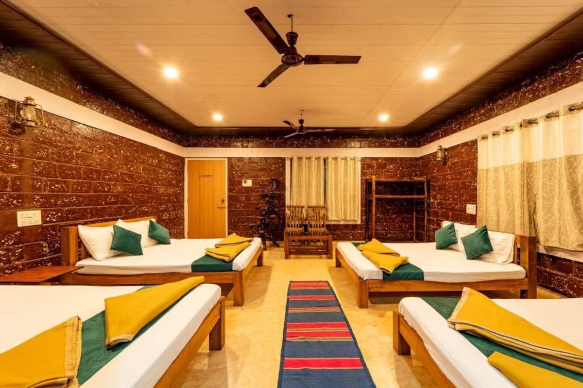 Ein Zimmer mit vier Einzelbetten, einem Deckenventilator, einer Holzbank, einem farbenfrohen Teppich und einer Ziegelwand.