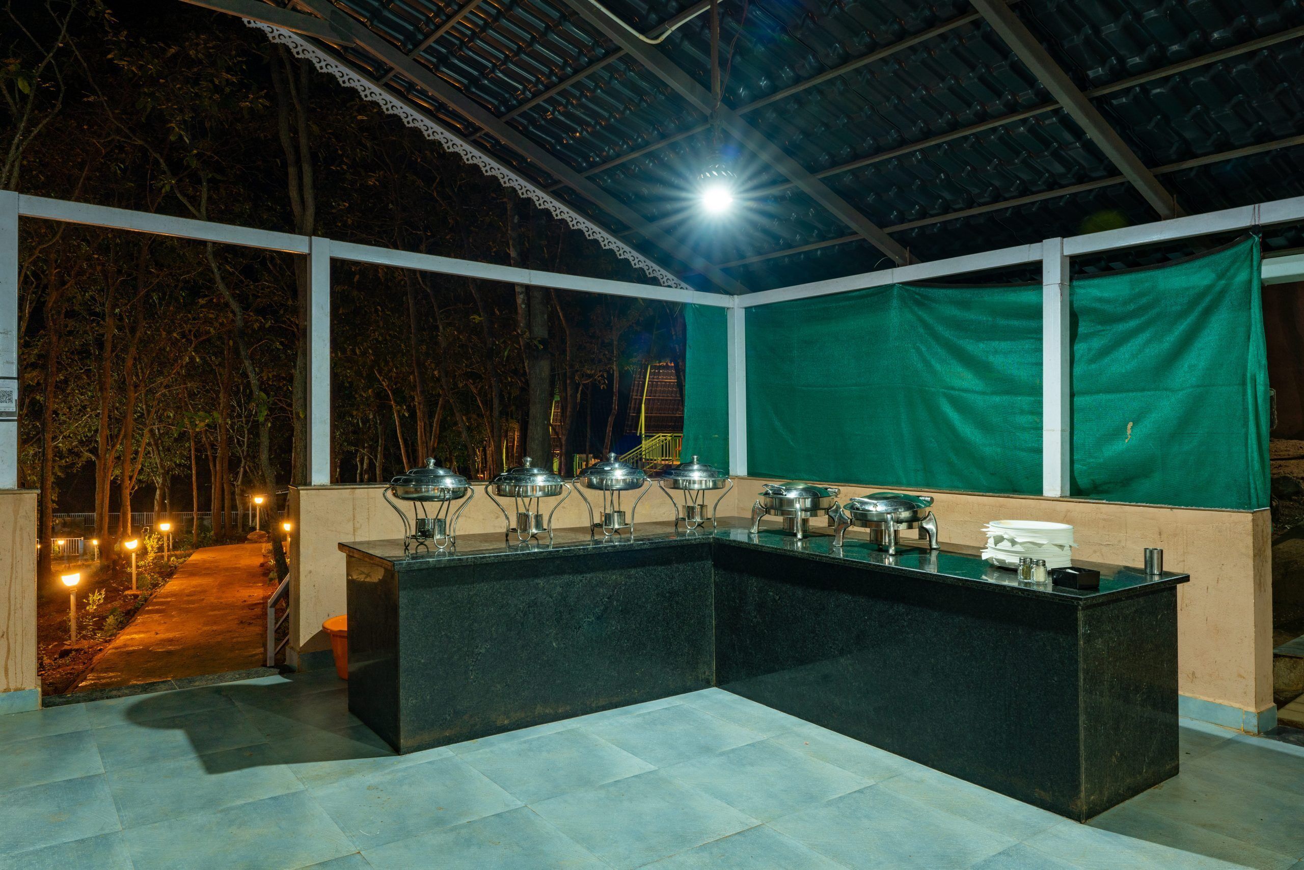 An outdoor bar area with a counter and multiple serving stations, surrounded by a covered patio with a view of trees and a pathway.