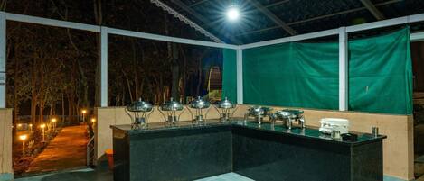 An outdoor bar area with a counter and multiple serving stations, surrounded by a covered patio with a view of trees and a pathway.