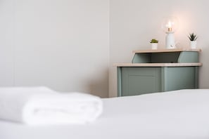 Cama bem arrumada com edredom branco, mesa de cabeceira com abajur e duas pequenas plantas em vasos.