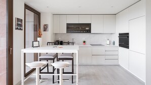 Una cocina moderna con muebles blancos, una isla central con taburetes, horno empotrado y encimera con hervidor y una pequeña planta.