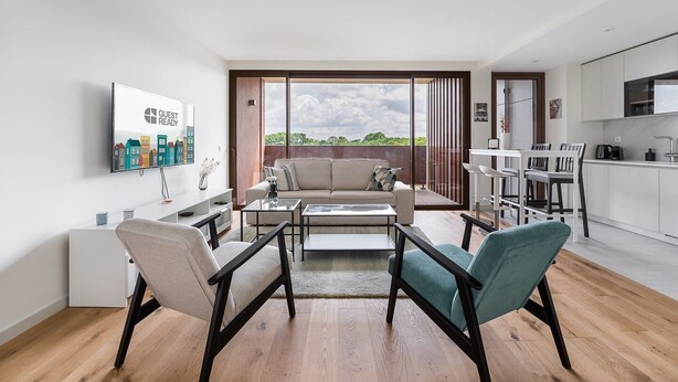 Una sala moderna con un sofá, dos sillones, una mesa de centro y un televisor montado en la pared. Hay un balcón con vistas a zonas verdes y un paisaje urbano.
