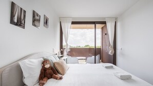 Una habitación de hotel con una cama, un peluche de mono, una ventana con cortinas y un balcón con mobiliario exterior.