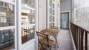 Ein schmaler Balkon mit Holztisch und -stühlen, großen Fenstern und Blick auf ein Gebäude mit Balkonen.