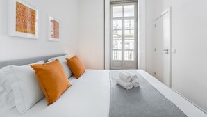 Ein modernes Hotelzimmer mit einem großen Bett, zwei orangefarbenen Kissen, einem grauen Teppich und einem Fenster mit Blick auf Gebäude.