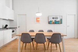 Una cocina moderna con armarios blancos, microondas y cafetera. Hay una mesa de comedor con cuatro sillas y una lámpara colgante encima. En la pared, un cuadro con el texto "GUEST READY".